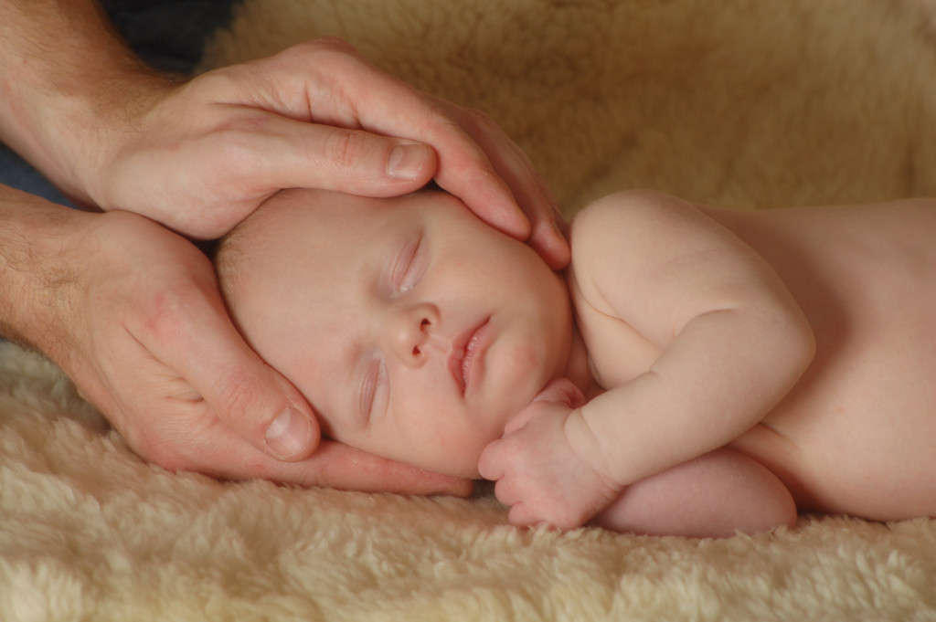 An image shows hands holding a lying baby by the baby's head.