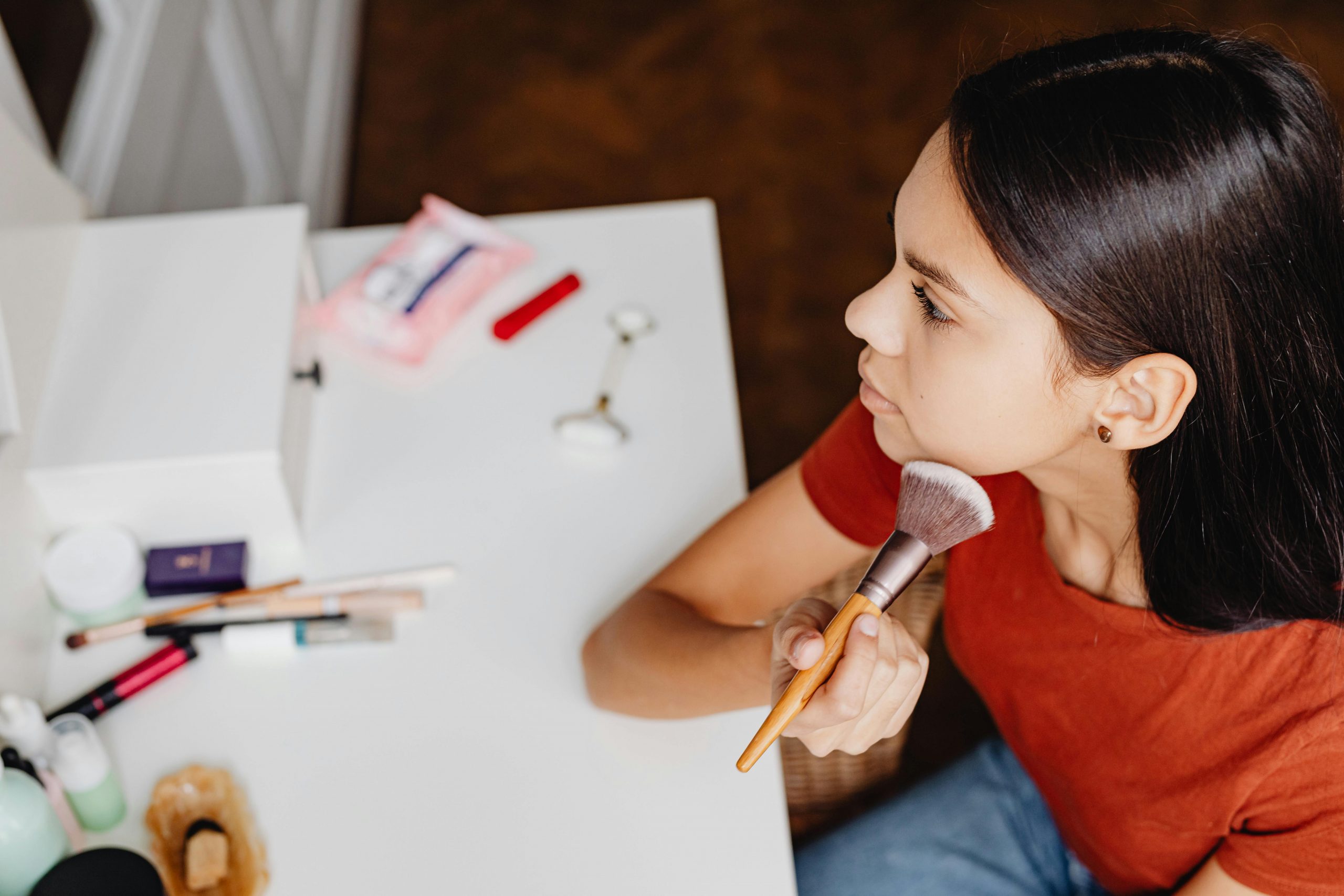 woman in a red shirt, looking in a mirror and applying foundation to her cheek with a makeup brush