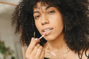 woman with curly hair, wearing a black tank top and applying a matte liquid lipstick to her lips with a wand