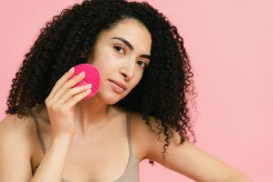 curly-haired woman in a gray tank top using a makeup sponge to apply makeup